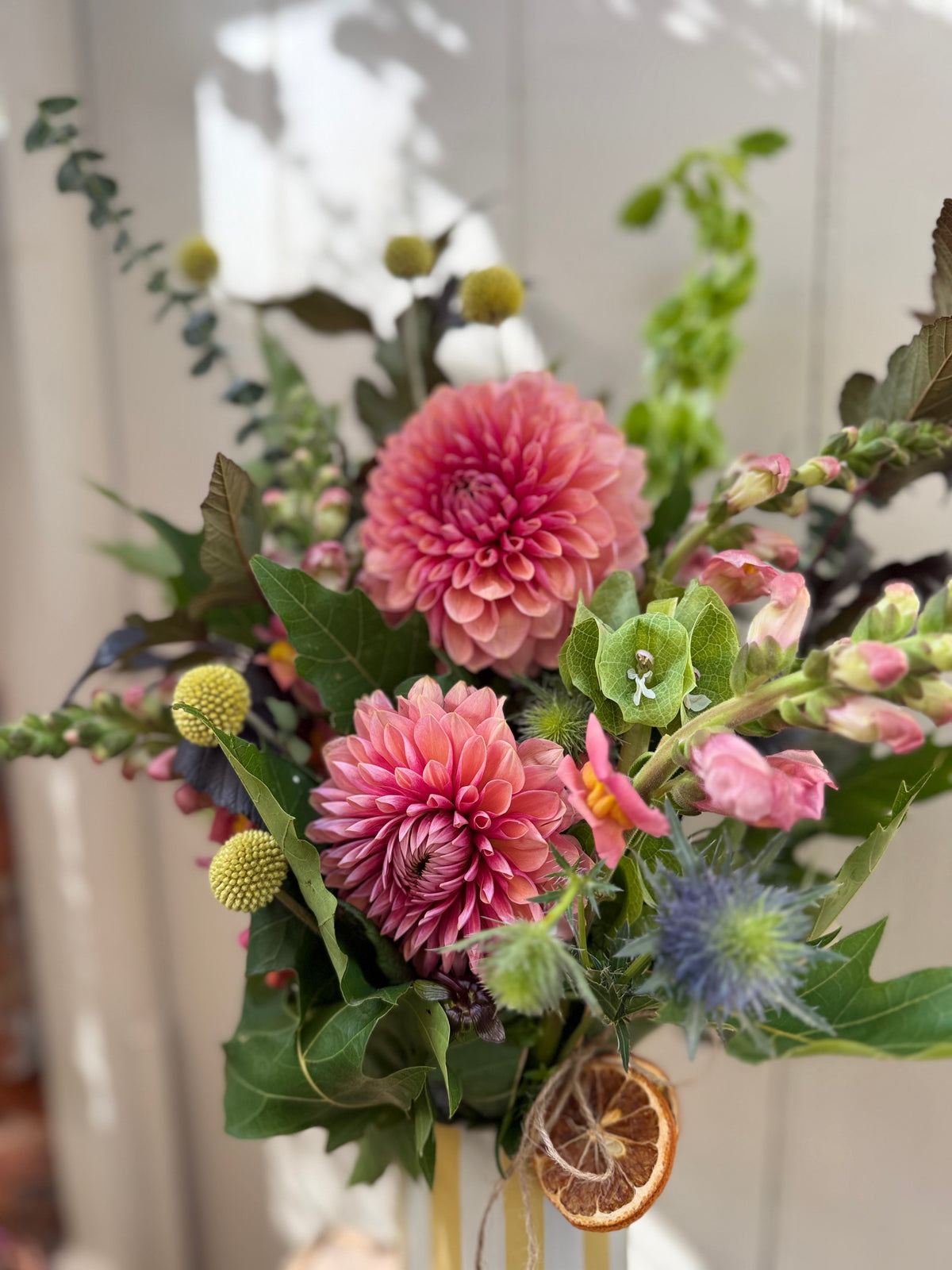 A close-up of a floral arrangement featuring two large pink dahlia flowers, green foliage, small yellow-green pom-pom flowers, a blue thistle-like bloom, and a dried orange slice hanging from the vase.